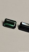 Photo showing two rectangular emerald-cut Paraiba colored tourmaline gemstones with teal blue and green hues on a plain background.