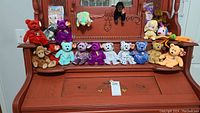 Wide photo showing collection of 15 Beanie Babies plush toys on piano with one package of heart protectors in the background.