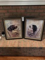 Two framed bird dog prints side by side on a brick hearth. Each print depicts a different dog with detailed backgrounds, wooden frames with gold trim.
