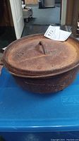 Cast iron pot with lid, marked size 10, sits on blue plastic container. Shows rust and wear; three feet visible at base.