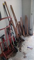 Various garden tools including rake, shovel, saws, axe with wooden handles arranged against wall showing condition and types.