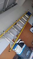 Photo showing full length of yellow Werner ladder leaning on wall above plastic containers on floor.