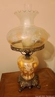 Full view of vintage carnival glass lamp showing marigold/peach globe with white floral pattern and brass base with claw feet.