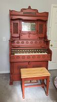 Full front view of red-painted antique pump organ with carved decoration, central replaced mirror, keyboard, stops and foot pedals, and yellow upholstered bench in front.