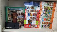 16 puzzle boxes stacked in a bookcase shelf showing various scenic locations and Disney World puzzle, piece counts from 300 to 1000 pieces