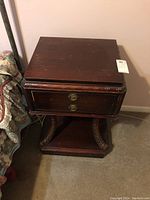 Front view of the dark wood vintage side table with two drawers and decorative brass handles, showing carved accents and open bottom shelf.