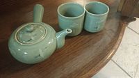 Tea set including teapot and two cups shown on wooden surface, jade green glaze with flying crane motif.