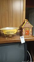 Both items on a wooden surface, showing the Fire King Pyrex casserole on warming stand next to the gingerbread house cookie jar