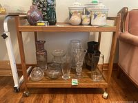 Front view of manufactured wood bar cart containing various glass and ceramic vases on two shelves, showing wood grain and caster wheels.