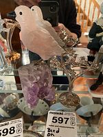 Rose quartz bird figurine perched on a purple amethyst crystal cluster with a decorative metal tree in the background.