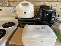 Four small appliances on kitchen counter: GE white hand mixer with beaters, Bosch black Tassimo coffee maker, Toastess white Panini maker, Starfrit black grill.