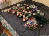 Overview of various Christmas tree ornaments including balls, drums, animals, and a strand of miniature tree lights on a dark comforter.
