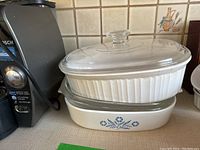 Two ceramic Corningware baking dishes stacked with clear Pyrex glass lids. Bottom dish has blue Cornflower pattern, top dish is white ribbed design (French White).