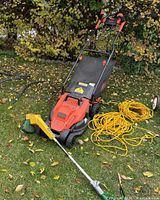 Black & Decker electric lawn mower with foldable handle, long yellow extension cords coiled, Weed Eater trimmer resting on ground, autumn foliage in background.
