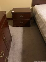 Photo of one wooden end table next to a bed, showing two drawers with metal pull handles and dark wood finish.