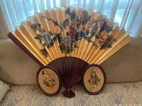 Photograph of the large decorative wall fan with floral print and two oval plaques with relief figures placed on a couch.