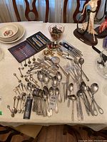 Overall view of assorted flatware including Sant Andrea stainless spoons, forks, knives, souvenir spoons, silver plated napkin rings, and other items on a table.
