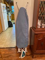 Ironing board in folded upright position covered with gray fabric protector, stands against wall.