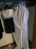 Two ladies evening dresses hanging on black hangers, one short black strapless with white overlay, one long light dress covered in a garment bag with visible buttons.
