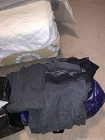 Photo showing a pile of men's clothing including several grey sweaters folded in a purple zippered tote bag on carpeted floor.