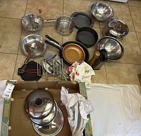 Overview of the kitchenware lot showing multiple pots, pans, colanders, cake pans, a sieve with a crank, and linens all on a tiled floor.