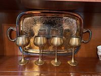 Four silver plated engraved goblets in front of a large silver plated engraved tray with handles, arranged on a wooden surface.