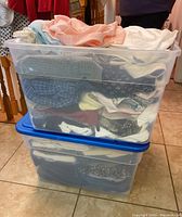 Two large plastic bins stacked with assorted women’s clothing visible inside. The clothing includes a variety of fabrics and colors, folded neatly inside the bins.