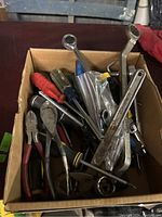 Box filled with assorted hand tools including screwdrivers, wrenches, and pliers showing wear and used condition