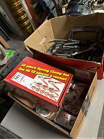 Two cardboard boxes, one holding a red box marked '12 piece Spring Clamp Set' and various clamps as well as miscellaneous hardware underneath. The second box contains orange straps, metal hooks, and more hardware parts.