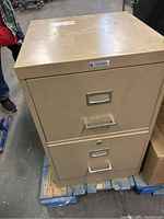 Front view of beige two drawer metal filing cabinet on wooden pallet showing wear and scuffs.