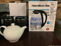 Hamilton Beach kettle in box with two white porcelain teapots and two Canon camera lens mugs in background