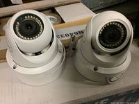 Two white dome HD security cameras side-by-side on cardboard, showing front with lenses and infrared LED rings.