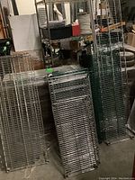 Multiple disassembled silver metal wire shelving rack pieces stacked vertically on the floor in a storage room environment with other equipment and boxes visible.