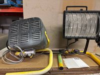 Side view of the black room heater with yellow stand, ventilation slots visible, electrical cord attached. The heater body is dusty and shows signs of use.