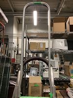 Front view of Magliner heavy duty hand dolly showing tall aluminum frame with green crossbars and shelving with boxes in background.