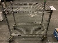Full view of the Metro metal wire rolling cart showing two wire shelves and four caster wheels on a concrete floor.