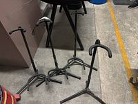 Four metal black string instrument stands arranged on floor, showing overall lot. Two stands missing parts.