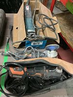 Photo of Ridgid angle grinder in bag with cords, Craftsman sander, and Makita drill in box on table