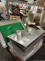 Metal trays stacked with green cutting boards and metal canisters on top amid other kitchen equipment.