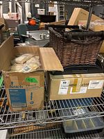 Photo of several cardboard boxes on metal shelving containing packaged wooden skewers and a dark woven wicker basket on top.