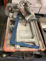 Top-down view of the Ridgid tile cutter with electric saw blade and blue angle ruler on top showing wear and dust.