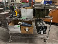 Full view of wire rack with three shelves containing various boxes and equipment. Metal frame and wire shelves are visible along with four swivel casters.