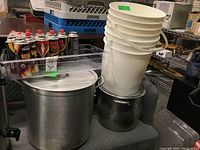 Wide view showing two stainless steel stock pots, stacked white plastic pails, and multiple butane fuel cartridges in a clear plastic container