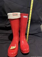 Front view of tall glossy red Hunter rain boots with white fleece liners, showing approximate height with measuring tape.