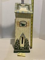 Front view of tall cream-colored wooden butterfly house with decorative bird painting and floral designs, measuring tape beside for scale showing 14 inches height.