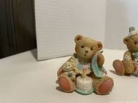 Cherished Teddy figurine holding a cake and baby doll, sitting down, pastel colors, with detailed textured fur.