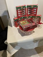 Multiple boxed Christmas ornaments in red and green, stacked on and around a white container decorated with snowflakes, along with a white decorative bell with red ribbon.