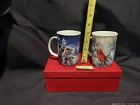 Two Christmas-themed ceramic mugs on top of closed red box with ruler showing about 4 inches height, black background.