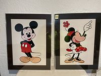 Pair of Disney framed glass wall art showing Mickey Mouse and Minnie Mouse standing and smiling, both in colorful classic outfits, propped against a white wall.