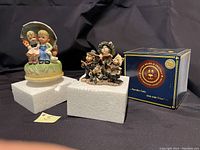Two figurines displayed side by side on white foam blocks with a dark background, including the Boyds Bears Sneaky Cats music box with its original blue box.